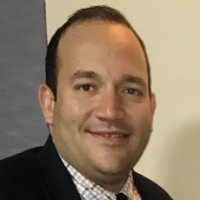 A profile of a smiling man with brown hair wearing a black suit jacket and plaid button-up shirt.